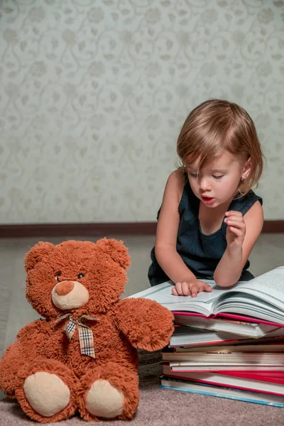 Bambina in abito blu scuro libro lettura seduta sul pavimento vicino orsacchiotto. Bambino legge storia per giocattolo . — Foto Stock