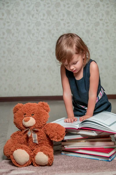 Fetiță în rochie albastră închisă citind o carte așezată pe podea lângă ursulețul de pluș. Copilul citește povestea pentru jucărie . — Fotografie, imagine de stoc