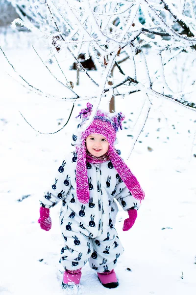 Enfant par temps neigeux. Bébé fille en suite blanche et chapeau rose, bottes gants dans le parc d'hiver de la neige. Heureux . — Photo