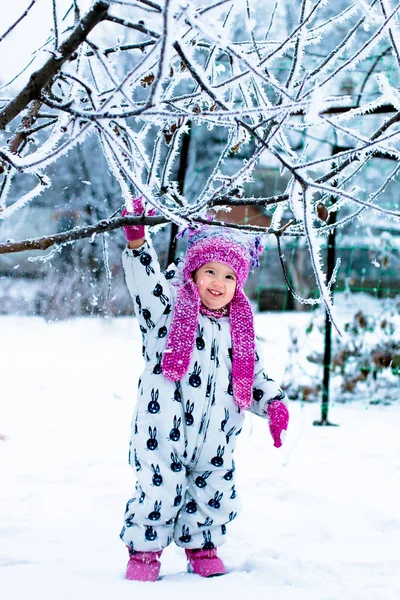 Enfant par temps neigeux. Bébé fille en suite blanche et chapeau rose, bottes gants dans le parc d'hiver de la neige. Heureux . — Photo