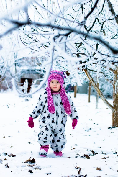 Enfant par temps neigeux. Bébé fille en blanc snowsuite et chapeau rose, bottes gants dans le parc d'hiver de neige . — Photo