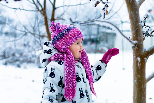 Enfant par temps neigeux. Bébé fille en blanc snowsuite et chapeau rose, bottes gants dans le parc d'hiver de neige . — Photo