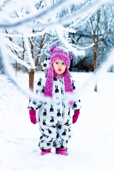 Enfant par temps neigeux. Bébé fille en blanc snowsuite et chapeau rose, bottes gants dans le parc d'hiver de neige . — Photo