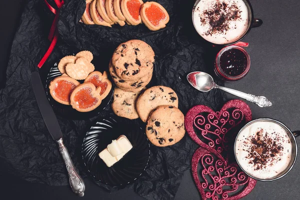 Romantik kahvaltı. İki bardak kahve, cappuccino çikolatalı kurabiye ve bisküvi siyah tablo arka plan üzerinde kırmızı kalpler yakınındaki ile. Sevgililer günü. Seviyorum. Üstten Görünüm. — Stok fotoğraf