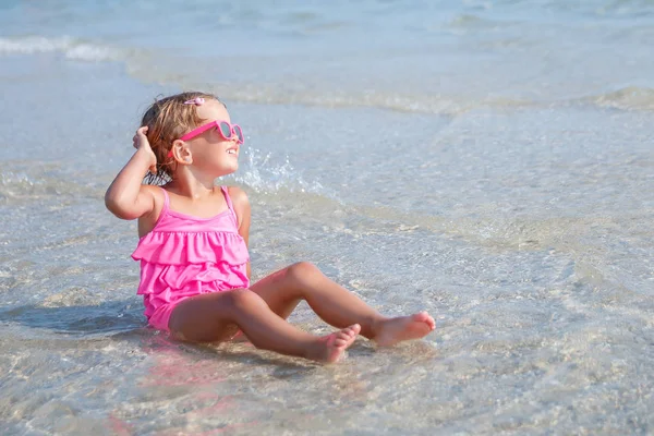 Παιδί σε ροζ μαγιό και γυαλιά ηλίου που κάθεται στη θάλασσα. Χαρούμενος, χαμογελώντας. Καλοκαιρινές διακοπές. Απολαύστε ήλιο. — Φωτογραφία Αρχείου
