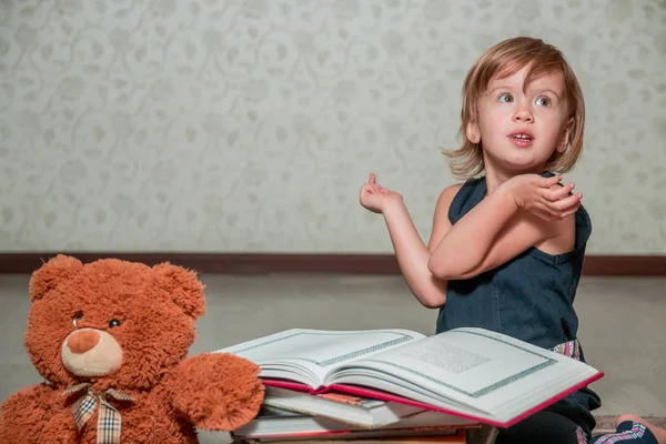 Una bambina vestita di blu scuro che legge un libro seduta sul pavimento vicino all'orsacchiotto. Bambino legge storia per giocattolo . — Foto Stock