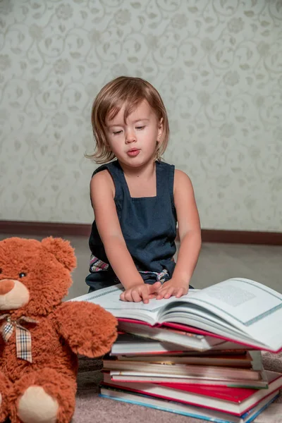 Una bambina vestita di blu scuro che legge un libro seduta sul pavimento vicino all'orsacchiotto. Bambino legge storia per giocattolo . — Foto Stock