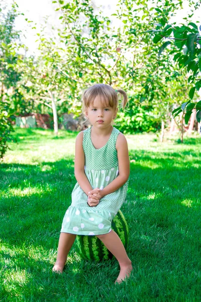 Niña en vestido verde sentada en la sandía en el parque, al aire libre. Verano. Niño triste. . —  Fotos de Stock