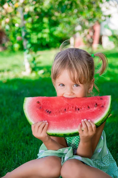 Liten flicka i grön klänning äta stor bit vattenmelon i parken. Sommartid. — Stockfoto
