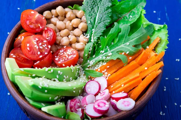 Vegan buddha bowl on blue wooden background. Bowl with carrot, lettuce, tomatoes cherry, radish, avocado and chickpea. Vegetarian, healthy, detox food concept. Close up.