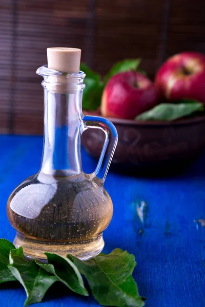 Cuka sari apel dalam botol kaca dengan latar belakang biru. Apel merah dalam mangkuk coklat . — Stok Foto