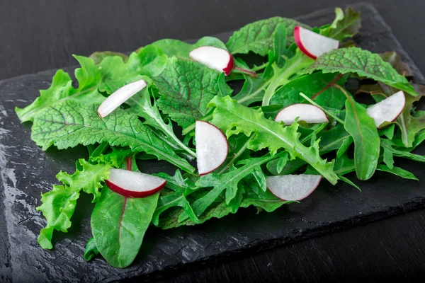 Gemischte Salatblätter mit Rettich auf schwarzem Schieferteller. Nahaufnahme. — Stockfoto