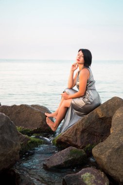 Kadın parlak gümüş elbisenin içinde taş deniz sahilde oturuyor.