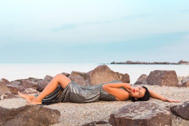 Kadın parlak gümüş elbisenin içinde taş deniz sahilde yatıyor.