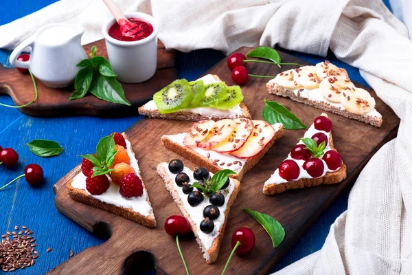 Tostadas de fruta sobre tabla de madera sobre fondo rústico azul. Desayuno saludable. Comer limpio. Concepto de dieta. Rebanadas de pan de grano con queso crema y varias frutas, bayas, semillas. Vegetariano. Vista superior . — Foto de Stock