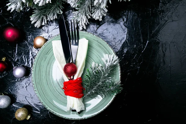Christmas table place setting. Holidays background. Green plate, knife and fork with christmas decorations on black table. Top view.
