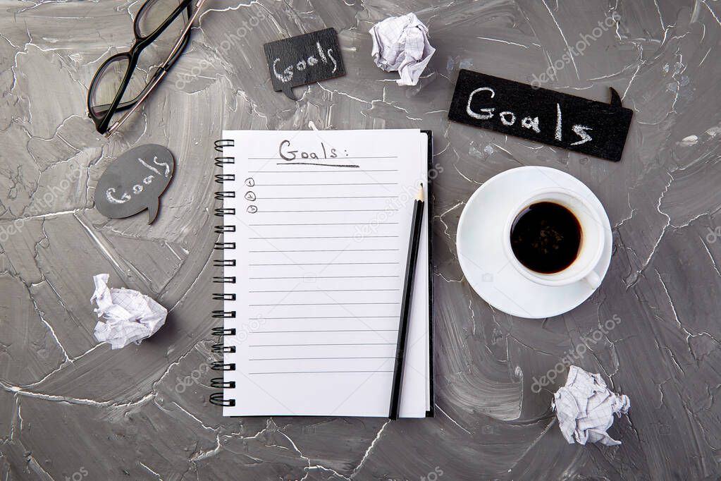 Goals as memo on notebook with idea, crumpled paper, cup of coffee over on grey background. Top view.