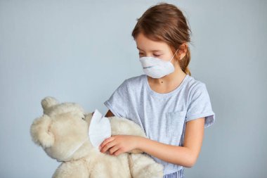Maskeli oyuncak ayıyı kucağına alan küçük bir kız. Coronavirus, covid-19 ve kirlilik koruma konsepti. Evde kal..