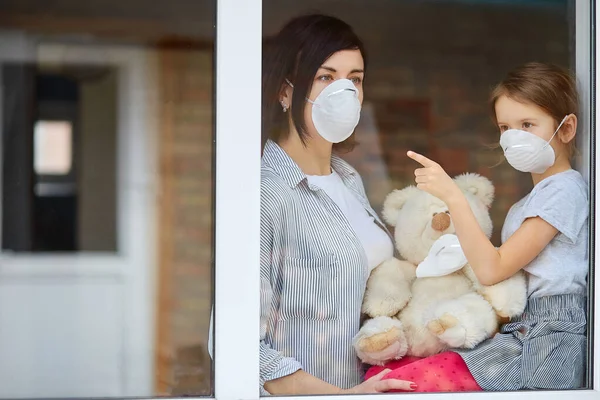 Moeder met dochter kind in masker kijken uit raam, coronavirus. — Stockfoto
