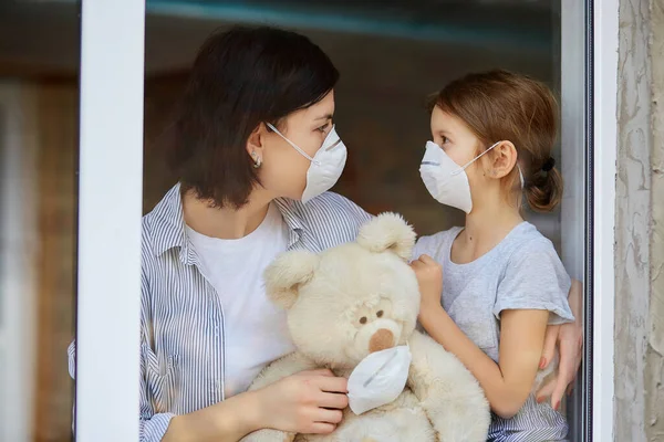 Moeder met dochter kind in masker kijken uit raam, coronavirus. — Stockfoto