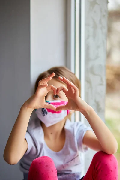 손으로 심장을 만드는 마스크를 쓴 어린 소녀, 코로나 바이러스 격리 — 스톡 사진