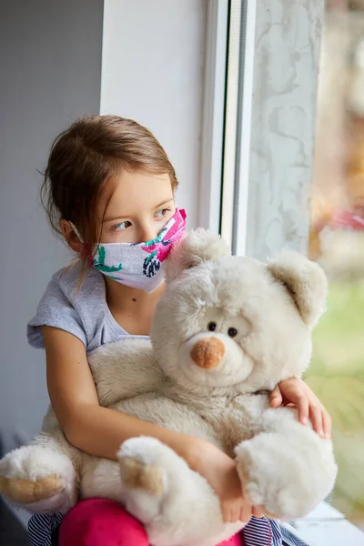 Klein meisje, kind in masker met teddybeer zit op ramen, coronavirus quarantaine — Stockfoto