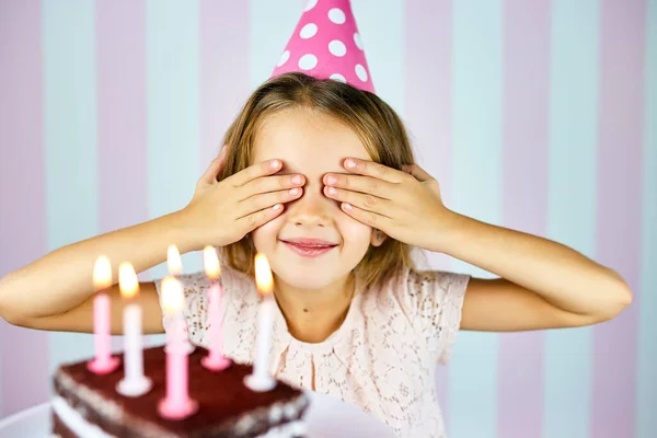 戴着粉色生日帽的金发小女孩笑着 闭上眼睛 许个愿 用蜡烛给巧克力生日蛋糕惊喜 孩子们庆祝她的生日 — 图库照片