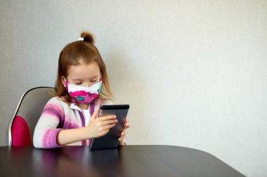 Evde koruyucu maske takan ve tablete oyun oynayan genç bir kız var. Coronavirus karantinası..