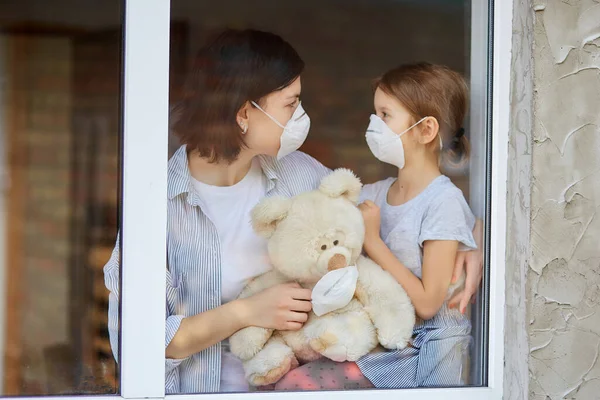 Mãe com filha criança em máscara olhando da janela, coronavírus . — Fotografia de Stock