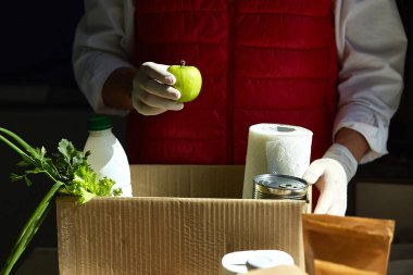 Koruyucu tıbbi maske ve eldiven giyen gönüllü bağış kutusuna yiyecek koyuyor. Kırmızı yelek giyen teslimatçı eleman yiyecek dolu bir kutuyla. Servis karantina pandemik koronavirüsü.