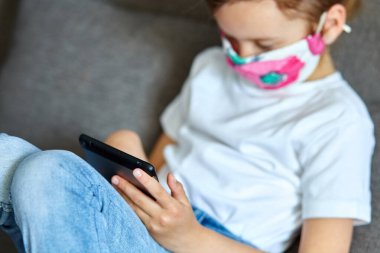 Korunma maskesi takan küçük kız tablet oynuyor ya da evdeki kanepede internetten video okuyarak öğreniyor. Evde kal. Coronavirus karantina ve sosyal uzaklık.