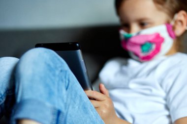 Korunma maskesi takan küçük kız tablet oynuyor ya da evdeki kanepede internetten video okuyarak öğreniyor. Evde kal. Coronavirus karantina ve sosyal uzaklık.