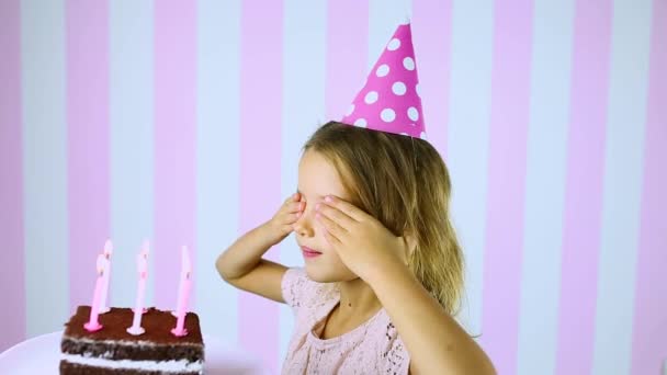 Szczęśliwa niespodzianka, zamknięte oczy dziewczynka w różowej czapce dmuchanie świece na torcie urodzinowym — Wideo stockowe