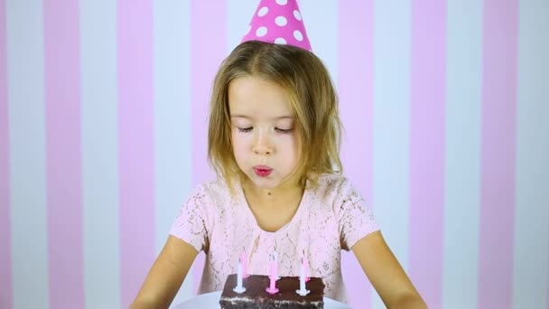 Menina feliz e sorridente em boné rosa soprando velas em um bolo de aniversário — Vídeo de Stock