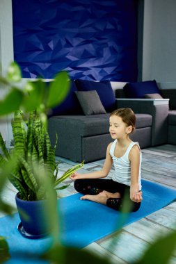 Mavi arka planda yoga sınıfında oturan bir kız. Evde eğitim, çocuk yogası, sakin ol, evde kal. Karantina, koronavirüs. Çocuklar evlerini karantinaya alırlar. Uluslararası yoga günü.
