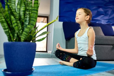 Küçük kız nilüfer çiçeğiyle meditasyon yapıyor, gözleri kapalı evde mavi mindere oturuyor, yoga yapıyor, dinleniyor. Karantina, koronavirüs. Çocuklar evlerini karantinaya alırlar. Uluslararası yoga günü.