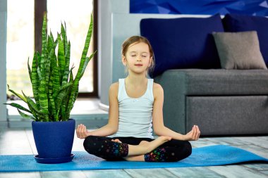 Küçük kız nilüfer çiçeğiyle meditasyon yapıyor, gözleri kapalı evde mavi mindere oturuyor, yoga yapıyor, dinleniyor. Karantina, koronavirüs. Çocuklar evlerini karantinaya alırlar. Uluslararası yoga günü.