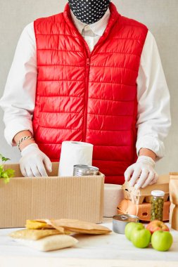 Koruyucu tıbbi maske ve eldiven giyen gönüllü bağış kutusuna yiyecek koyuyor. Kırmızı yelek giyen teslimatçı eleman yiyecek dolu bir kutuyla. Servis karantina pandemik koronavirüsü.