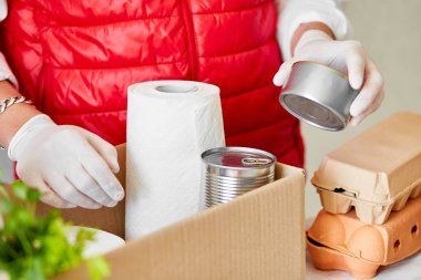 Koruyucu tıbbi maske ve eldiven giyen gönüllü bağış kutusuna yiyecek koyuyor. Kırmızı yelek giyen teslimatçı eleman yiyecek dolu bir kutuyla. Servis karantina pandemik koronavirüsü.