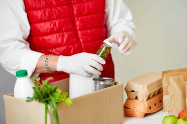 Koruyucu tıbbi maske ve eldiven giyen gönüllü bağış kutusuna yiyecek koyuyor. Kırmızı yelek giyen teslimatçı eleman yiyecek dolu bir kutuyla. Servis karantina pandemik koronavirüsü.