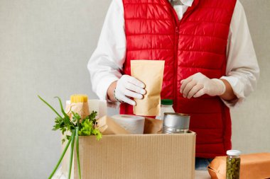 Koruyucu tıbbi maske ve eldiven giyen gönüllü bağış kutusuna yiyecek koyuyor. Kırmızı yelek giyen teslimatçı eleman yiyecek dolu bir kutuyla. Servis karantina pandemik koronavirüsü.