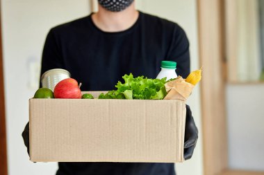 Siyah koruyucu maskeli ve eldivenli gönüllü eve bağış kutusu getiriyor. Paket kutusu yiyecek taşıyan kurye. Teslimatsız teslimat. Servis karantina pandemik koronavirüsü.