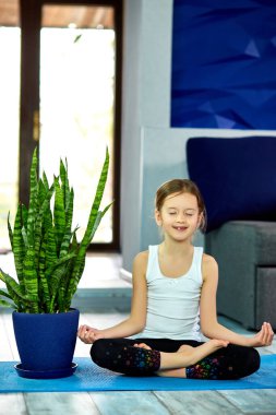Küçük kız nilüfer çiçeğiyle meditasyon yapıyor, gözleri kapalı evde mavi mindere oturuyor, yoga yapıyor, dinleniyor. Karantina, koronavirüs. Çocuklar evlerini karantinaya alırlar. Uluslararası yoga günü.
