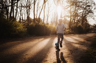 Küçük beyaz çocuk ormanda futbol oynuyor.