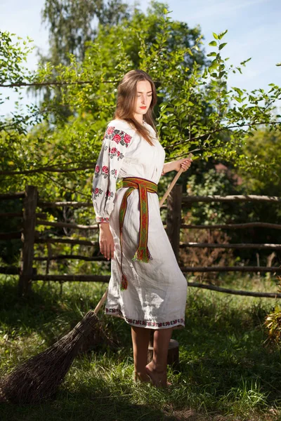 草の上の夏の家の外の木製仏舎利塔の横にほうきと国民服の長い髪の美しいウクライナの女の子刺繍 — ストック写真