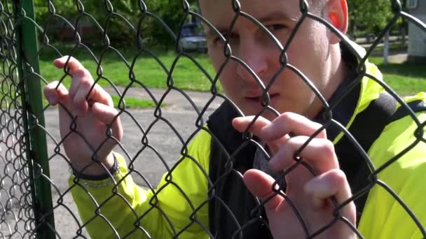 Un hombre de unos 20 años, apoyado contra la valla mirando a la cámara — Vídeo de stock