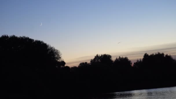 Uma lua crescente sobre o lago ao entardecer, com silhueta, floresta e avião voador na distância ao fundo — Vídeo de Stock