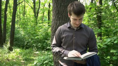 Genç işadamı ormanda bir ağacın yanında kitap okuyor.