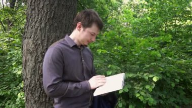 Genç işadamı ormanda bir ağacın yanında kitap okuyor.