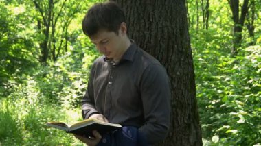 Genç işadamı ormanda bir ağacın yanında kitap okuyor.